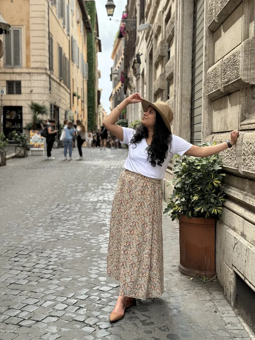 Arsha in a sun hat posing against a warm stone Roman wall beside a flowerpot