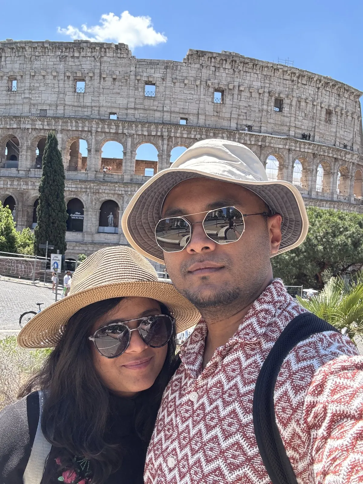 Arsha and Binu in sun hats and sunglasses taking a selfie in front of the Colosseum under a clear blue sky