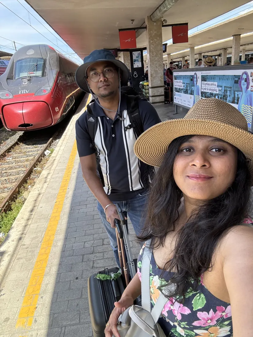 Selfie of Arsha in a sun hat and Binu on the platform at Florence, a red high-speed train behind them