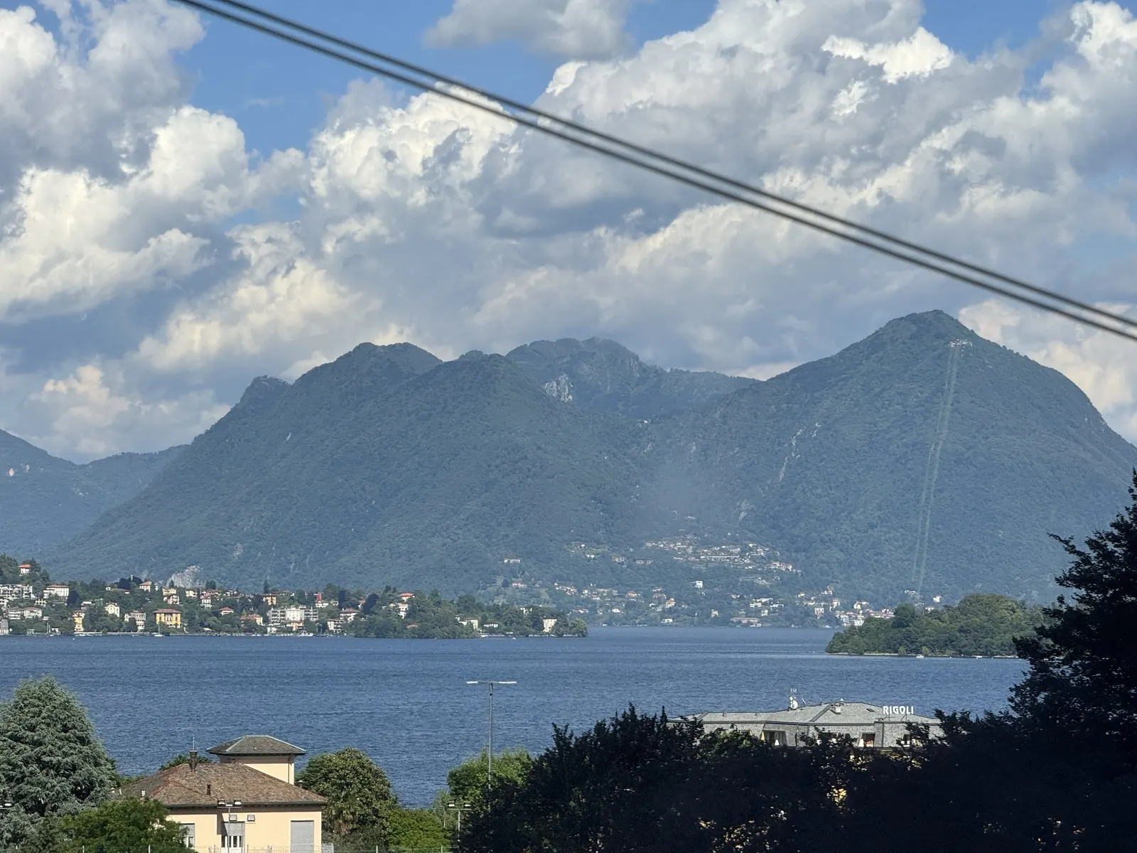 Lake Maggiore from the train window: blue water, a small town on the shore and green mountains under big white clouds