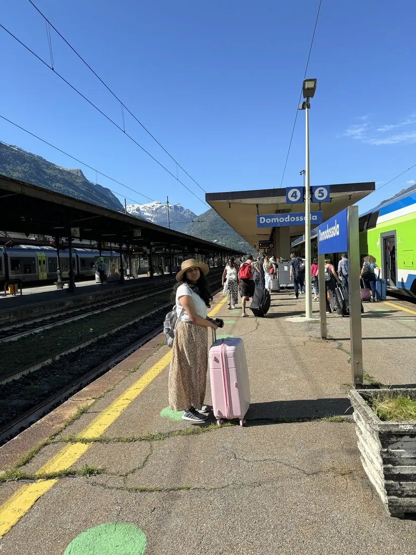 Arsha with a pink suitcase on the sunny platform at Domodossola, mountains behind