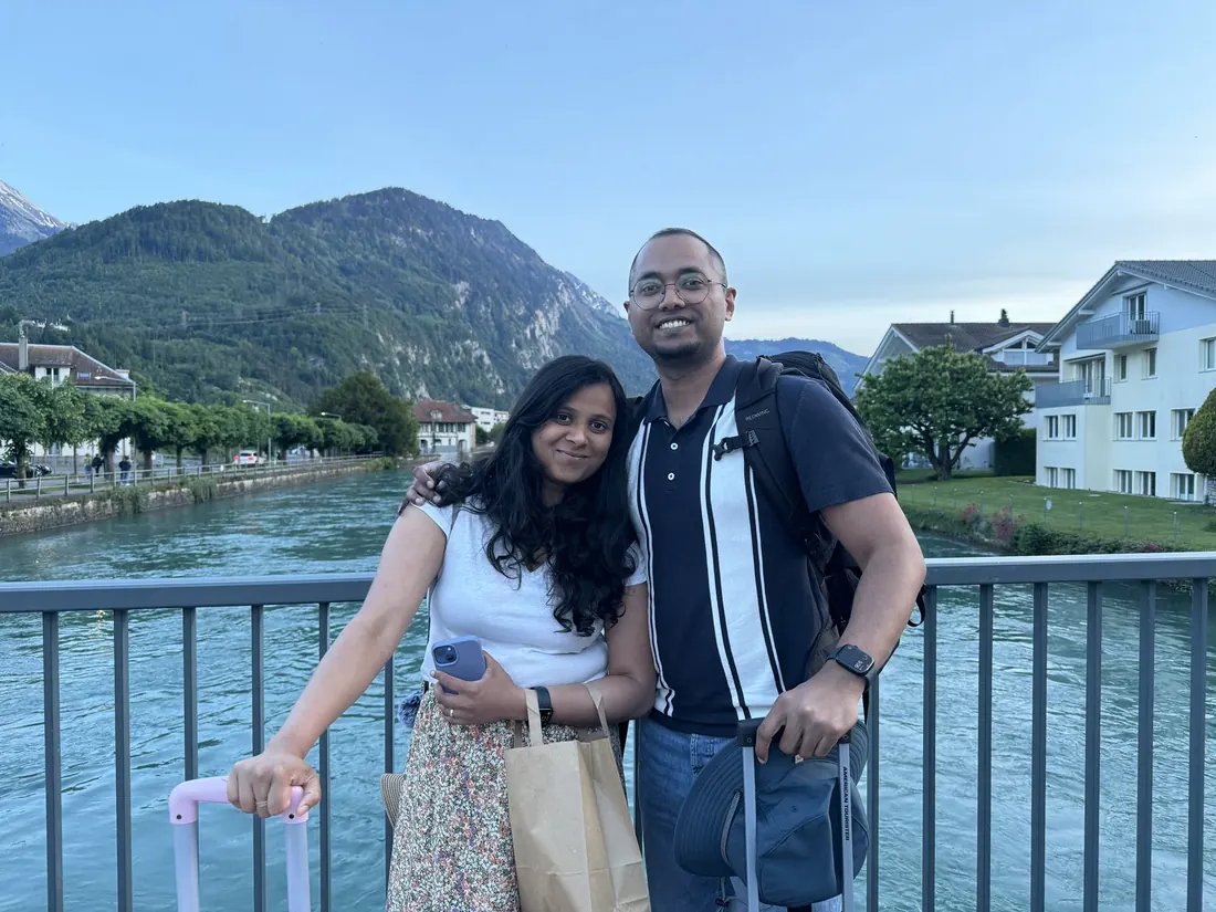 Arsha and Binu on the bridge over the turquoise Aare in Interlaken at dusk, the pink suitcase beside them