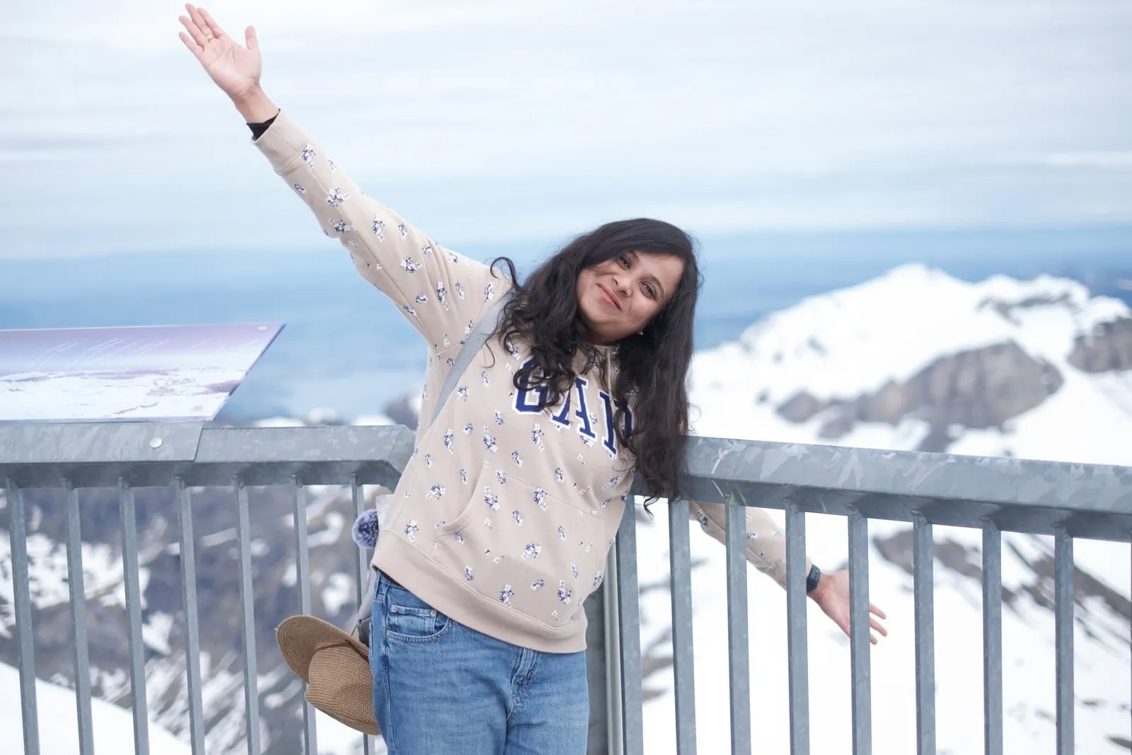 Arsha with one arm raised at the Schilthorn viewpoint railing, snowy peaks behind her