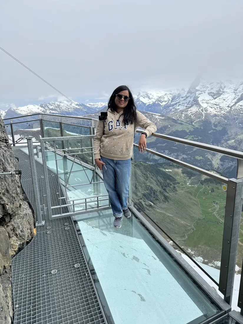 Arsha standing on the glass floor of the Birg Thrill Walk, a cliff edge and mountains behind