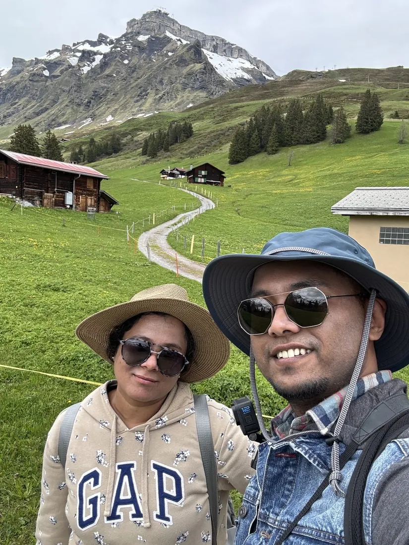 Arsha and Binu in sun hats taking a selfie on the trail, a chalet and peaks behind