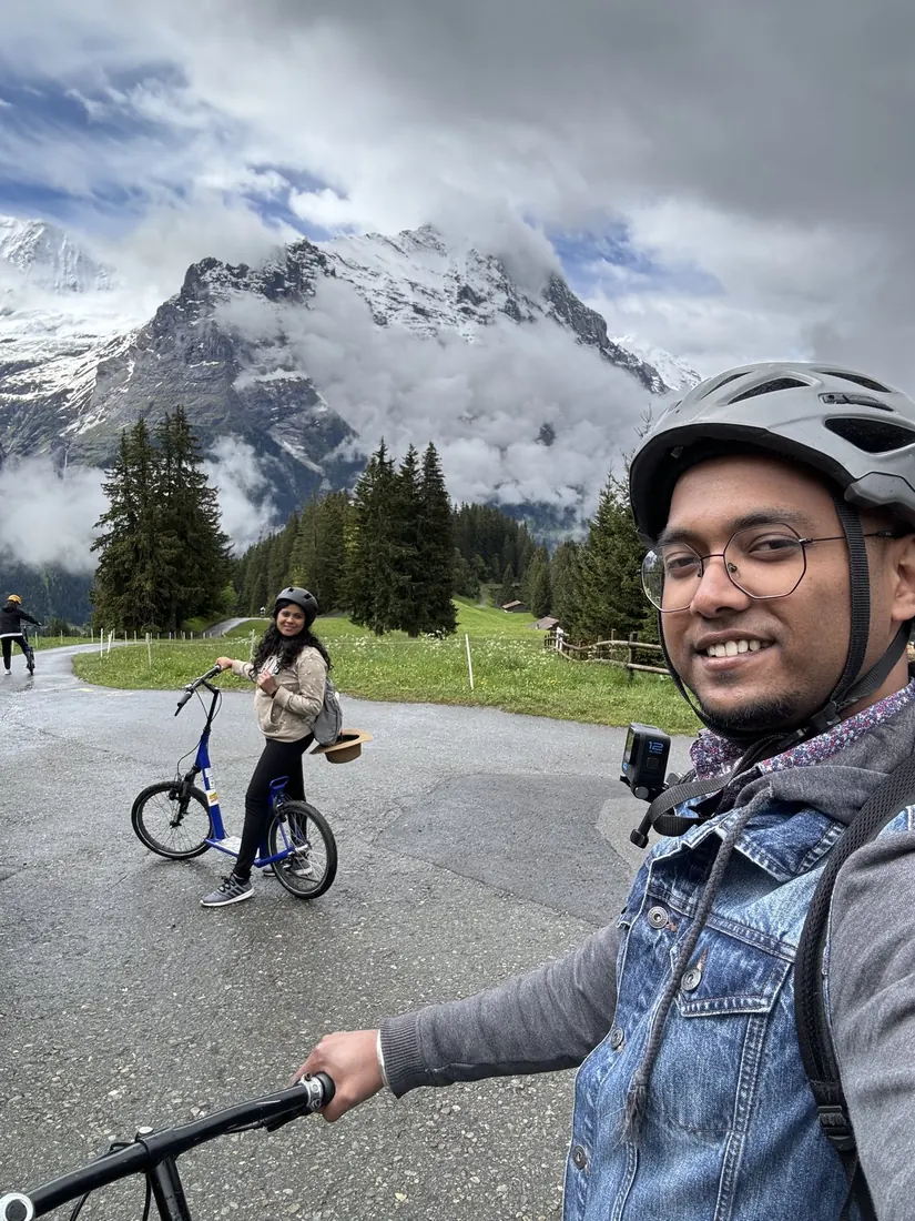 Binu in a helmet taking a selfie with Arsha riding a Trottibike behind him, snowy peaks in the distance