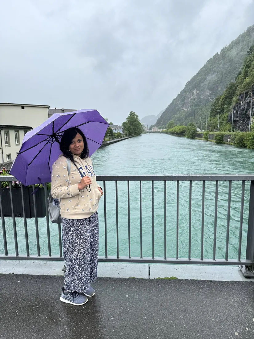 Arsha under a purple umbrella on a bridge over the turquoise Aare, mountains in cloud behind