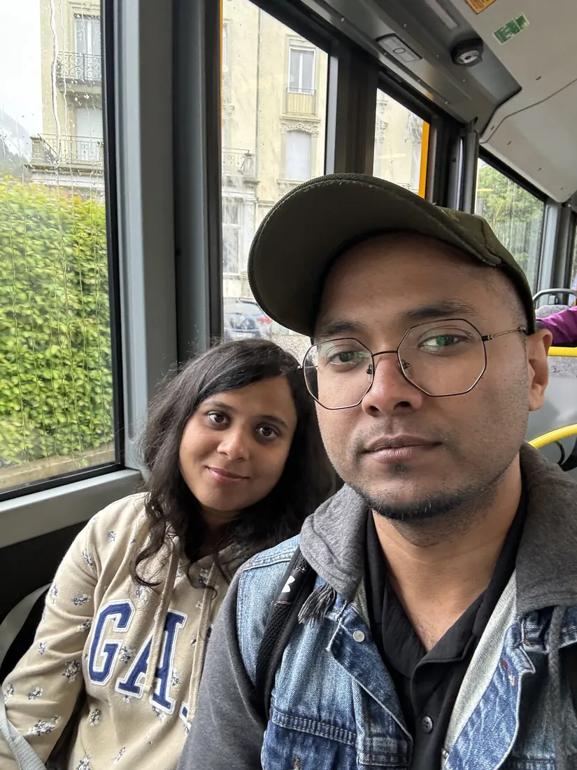Arsha and Binu in hoodies taking a selfie on a city bus, rain on the windows