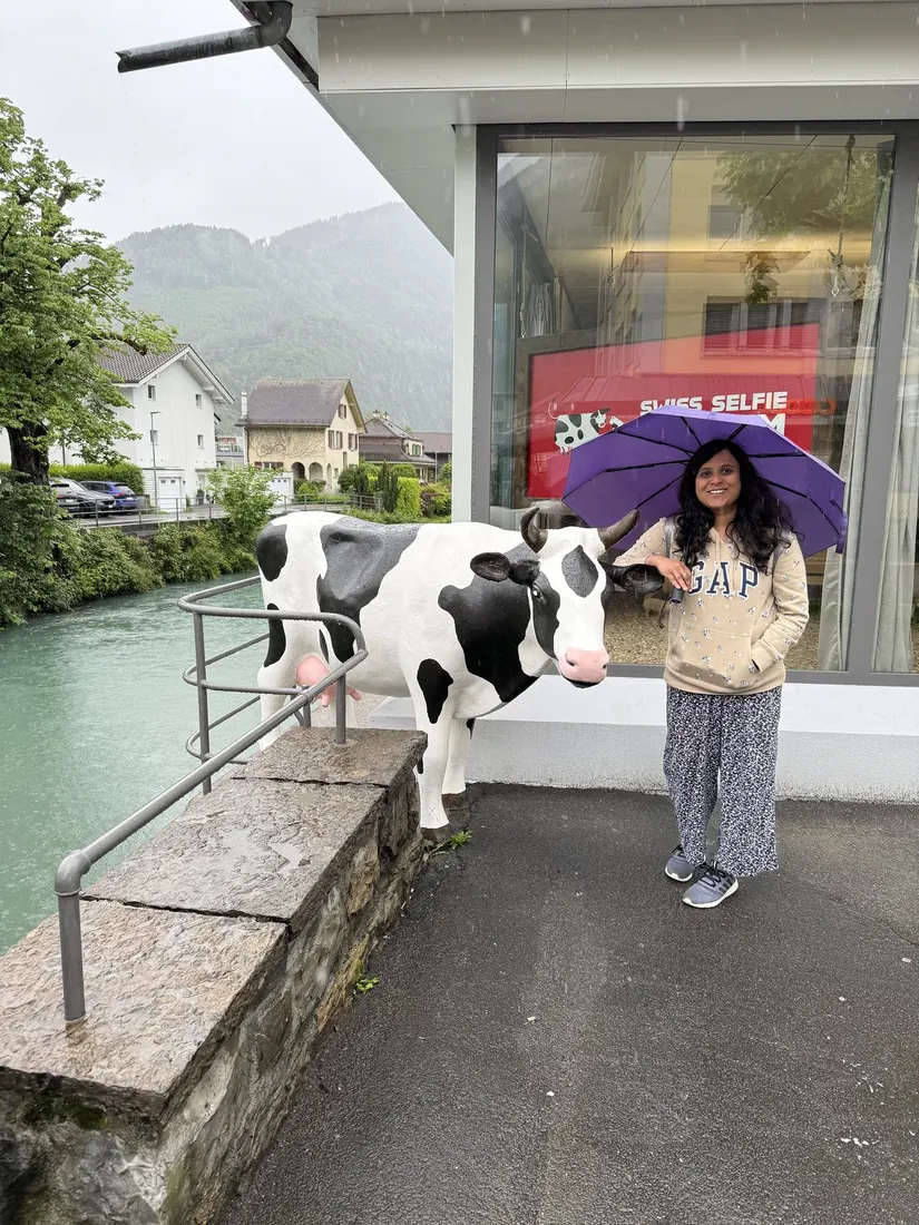 Arsha with a purple umbrella next to a life-size black-and-white cow statue on a river bridge