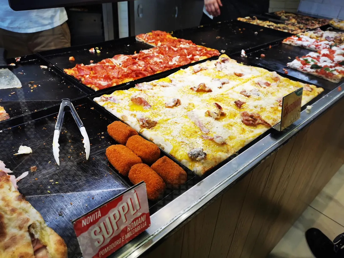 Rows of golden supplì beside pizza slices in a takeaway counter, under a sign reading Supplì Romani